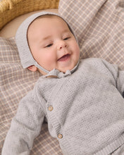 Load image into Gallery viewer, A baby wearing a light gray knit bonnet and matching long-sleeve cardigan lies on a beige and white plaid blanket.
