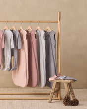 Load image into Gallery viewer, A set of baby sleep sacks and long-sleeve bodysuits in soft pastel colors including pink, dusty rose, light gray, and charcoal, hanging on a wooden clothing rack with a small rustic stool in the foreground.

