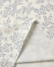 Load image into Gallery viewer, A close-up of a folded fabric item, likely a blanket or throw, featuring a light gray botanical leaf pattern on an off-white background, with a folded corner showing a textured cream lining.
