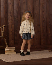 Load image into Gallery viewer, A young girl with curly hair stands in front of a dark wood plank wall wearing a cream knit cardigan with black star patterns, a dark patterned pleated skirt, white socks, and black shoes.
