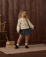 Load image into Gallery viewer, A young girl with curly blonde hair stands in a rustic room with dark wood paneling, wearing a cream cardigan with black star patterns, a dark blue patterned pleated skirt, white socks, and black shoes.
