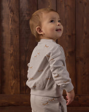 Load image into Gallery viewer, A toddler standing in profile against a dark wood plank wall, wearing a light beige long-sleeve top and matching pants with small tan embroidered motifs.
