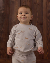 Load image into Gallery viewer, A smiling toddler wearing a light beige long-sleeve sweatshirt and matching sweatpants with a small brown fox print, standing against a dark wood plank background.
