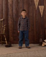 Load image into Gallery viewer, A young boy stands in front of a rustic dark wood plank wall wearing a long-sleeve plaid button-up shirt in muted red and blue tones, paired with blue jeans and brown boots. A wooden chair, pinecone, lantern, and small banner decorations are visible in the setting.
