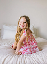 Load image into Gallery viewer, Young girl in pink horseshoe-patterned pajamas sitting on a bed.
