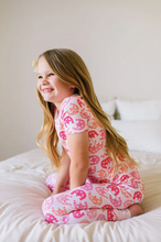 Load image into Gallery viewer, Young girl wearing pink horseshoe-patterned pajamas sitting on a white bed.
