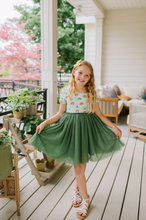 Load image into Gallery viewer, Young girl in a green skirt and floral top standing on a wooden deck with plants and furniture around.
