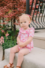 Load image into Gallery viewer, Young child in a pink romper sitting on a stone ledge with flowers and a bench in the background.
