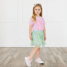 Load image into Gallery viewer, Young girl wearing a pink top with a bow and a green skirt on a wooden floor.
