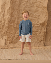 Load image into Gallery viewer, Child wearing a blue long-sleeve shirt and patterned shorts standing against a brown textured backdrop.
