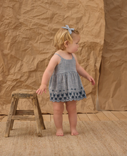 Load image into Gallery viewer, Child in a blue checkered dress standing next to a wooden stool against a brown paper backdrop.
