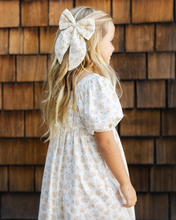 Load image into Gallery viewer, Young girl wearing a white dress with a large bow in her hair against a wooden wall.
