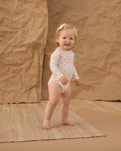 Load image into Gallery viewer, Child in a floral onesie standing on a textured rug with a brown backdrop
