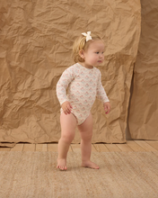 Load image into Gallery viewer, Child wearing a floral onesie standing against a textured brown backdrop
