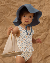 Load image into Gallery viewer, Baby in a swimsuit and blue sun hat holding a toy sailboat against a brown textured background
