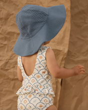 Load image into Gallery viewer, Child wearing a blue sun hat and patterned swimsuit against a brown background
