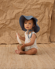 Load image into Gallery viewer, Baby sitting on a textured surface wearing a blue sun hat and a swimsuit against a brown paper bag background.
