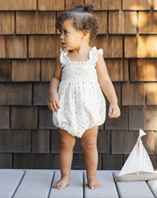 Load image into Gallery viewer, Child in a floral romper standing on a wooden deck with a sailboat model.
