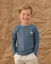 Load image into Gallery viewer, Child wearing a blue long-sleeve shirt with a logo against a brown textured background
