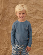 Load image into Gallery viewer, Young boy wearing a blue long-sleeve shirt with a logo against a textured brown background
