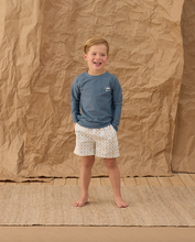 Load image into Gallery viewer, Child wearing a blue long-sleeve shirt and patterned shorts standing against a brown paper backdrop.
