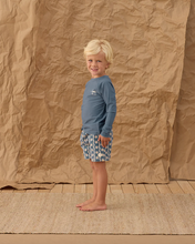 Load image into Gallery viewer, Young boy in blue shirt and patterned shorts standing against a textured brown backdrop
