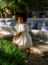 Load image into Gallery viewer, Child in a white dress standing in front of a decorative bench with mosaic patterns.
