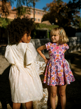 Load image into Gallery viewer, Two young girls in dresses standing outdoors with a building and trees in the background.
