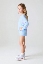 Load image into Gallery viewer, Young girl wearing a light blue outfit with colorful accents on a white background
