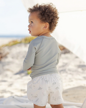 Load image into Gallery viewer, Child wearing a light gray long-sleeve shirt and white shorts with a pattern, standing on a sandy beach.

