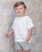 Load image into Gallery viewer, Child wearing a white t-shirt with small patterns and light gray shorts standing against a stone wall.
