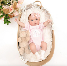 Load image into Gallery viewer, Baby in a pink onesie with bunny design, sitting in a decorative crib with flowers and a teddy bear.

