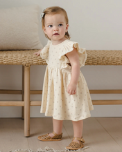 Load image into Gallery viewer, Young child wearing a light-colored dress with ruffled sleeves, standing in a room with a wooden bench.
