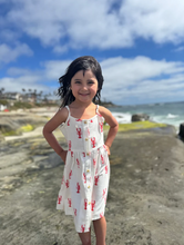 Load image into Gallery viewer, Young girl in a white dress with red patterns standing on a beach.
