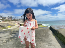 Load image into Gallery viewer, Young girl in a white dress with red lobster prints standing on a rocky beach.
