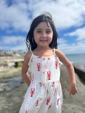 Load image into Gallery viewer, Young girl in a white dress with red lobster patterns standing on a beach.
