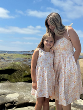 Load image into Gallery viewer, Two women in matching floral dresses standing on a rocky beach with a blue sky.
