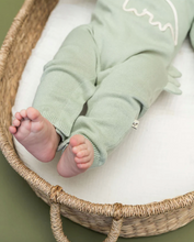 Load image into Gallery viewer, Baby's legs in light green pants inside a woven basket on a white surface.
