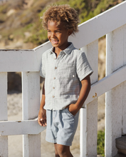 Load image into Gallery viewer, Child wearing a light blue shirt and shorts standing on a white wooden deck with a natural background.
