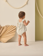 Load image into Gallery viewer, Baby in a floral romper standing in a minimalistic room with a wooden sculpture and green wall.

