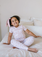 Load image into Gallery viewer, Child wearing pink pajamas with pink patterns sitting on a bed.
