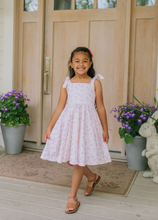 Load image into Gallery viewer, Young girl in a white floral dress standing on a wooden deck with potted plants and a wooden door in the background.
