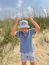 Load image into Gallery viewer, Child wearing a blue shirt and shorts with a hat, standing on sand with grasses in the background
