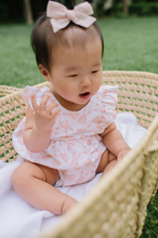 Load image into Gallery viewer, Baby in a floral outfit with a bow sitting outdoors on grass.
