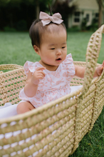Load image into Gallery viewer, Baby in a woven basket with a bow on head outdoors
