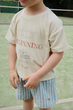 Load image into Gallery viewer, Child wearing a light-colored shirt with text and blue shorts on a tennis court.
