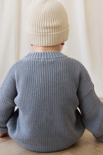 Load image into Gallery viewer, Child wearing a blue knitted sweater and beige hat sitting on a wooden floor.
