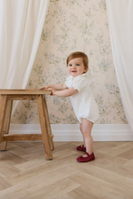 Load image into Gallery viewer, Child in a white dress standing next to a wooden stool against a floral wallpapered wall.
