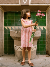 Load image into Gallery viewer, Young girl in a red floral dress holding flowers in front of a green tiled wall.
