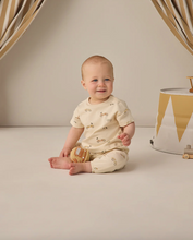 Load image into Gallery viewer, Baby in a yellow outfit sitting on a light gray floor with a neutral-colored wall and decorative elements in the background.
